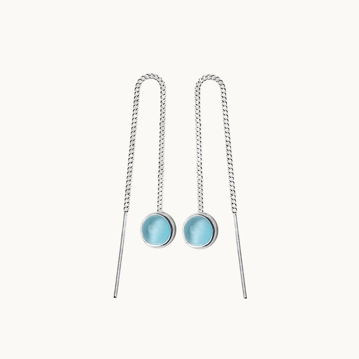 Une paire de boucles d'oreilles est exposée devant un fond beige.  Ce sont des boucles d'oreilles pendantes. Elles sont en chaine avec une pierre ronde opale bleue au bout. Elles sont en argent 925. 