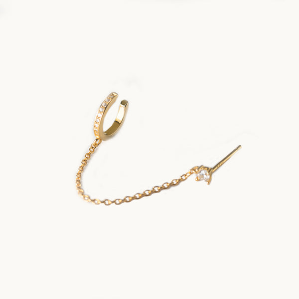 Une boucle d'oreille est exposée devant un fond beige.  C'est un diamant avec une chaîne et un anneau en strass qui se met sur le cartilage. La boucle est en plaqué or.  