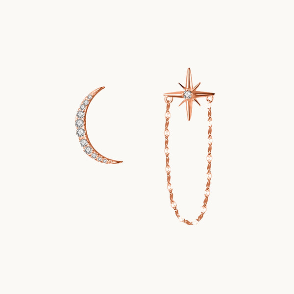 Une paire de boucles d'oreilles est exposée devant un fond beige.  Ce sont des boucles dépareillées. L'une est un croissant de lune orné de strass et l'autre est une étoile avec un strass au milieu et une chaîne pendante. Elles sont en argent 925 plaqué or rose. 