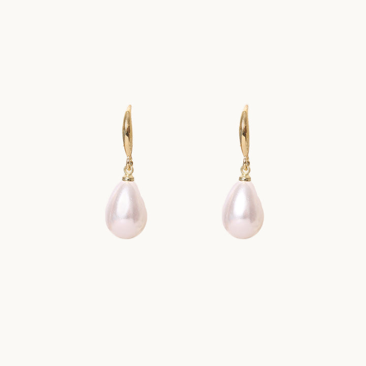 Une paire de boucles d'oreilles est exposée devant un fond beige.  Elles sont pendantes avec une tige dorée et une perle ovale. 