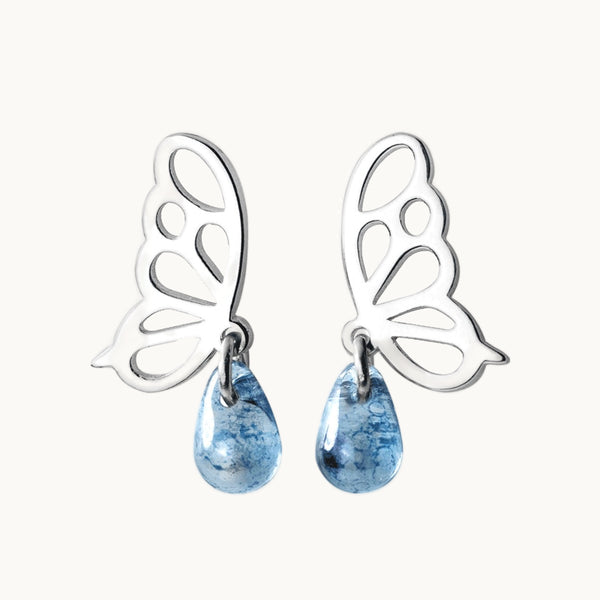 Une paire de boucles d'oreilles est exposée devant un fond beige.  Ce sont des boucles en forme de papillon de profil sur lesquel pend une goutte en pierre transparente bleue. Elles sont en argent 925. 