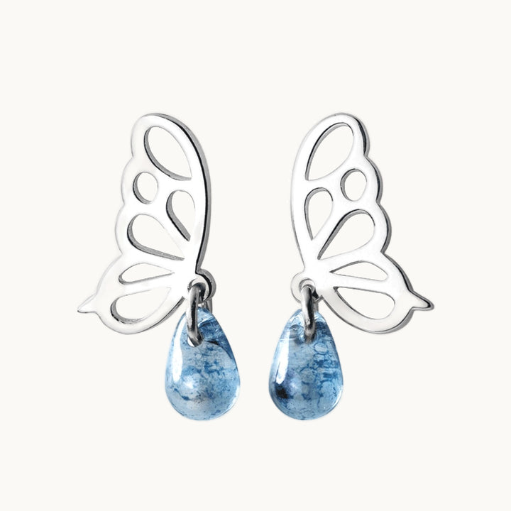 Une paire de boucles d'oreilles est exposée devant un fond beige.  Ce sont des boucles en forme de papillon de profil sur lesquel pend une goutte en pierre transparente bleue. Elles sont en argent 925. 