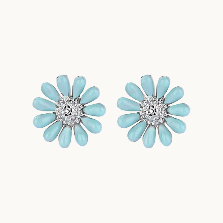 Une paire de boucles d'oreilles est exposée devant un fond beige.  Ce sont des fleurs aux pétales bleu ciel et au cœur en strass. Elles sont en argent 925. 