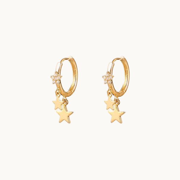 Une paire de boucles d'oreille est exposée devant un fond beige.  Ce sont des anneaux dorées sur lesquels pendent des étoiles. Il y a deux étoiles dorées et une étoile ornée de strass qui pendent sur l'anneau. 