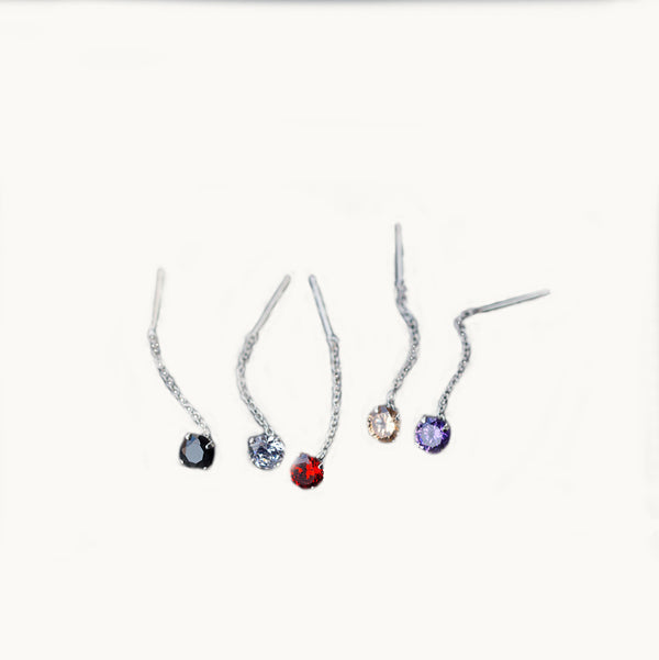 5 boucles d'oreilles sont exposées devant un fond beige.  Ce sont des boucles d'oreille longues. Il y a un diamant, disponibles en 5 couleur différentes, sur laquelle pend une chaîne en argent 925. 