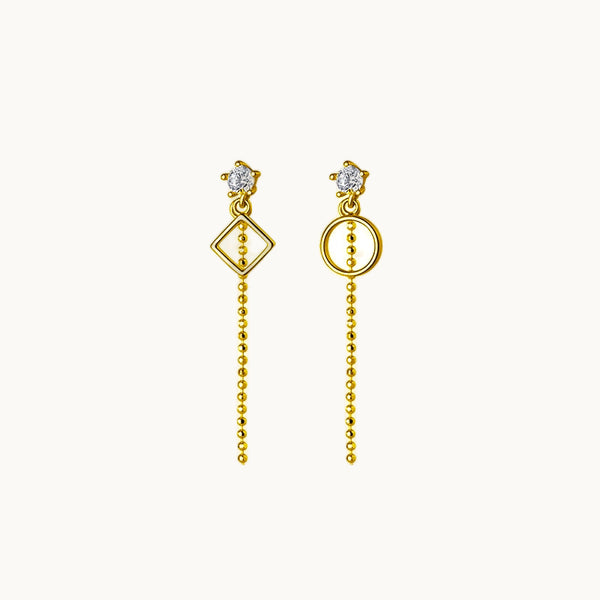 Une paire de boucles d'oreilles est exposée devant un fond beige.  Ces boucles sont composées d'un diamant sur lequel pend une chaîne et une forme géométrique qui n'est pas la même sur les deux boucles ; il y a un rond et un losange. Elles sont en argent 925 plaqué or.