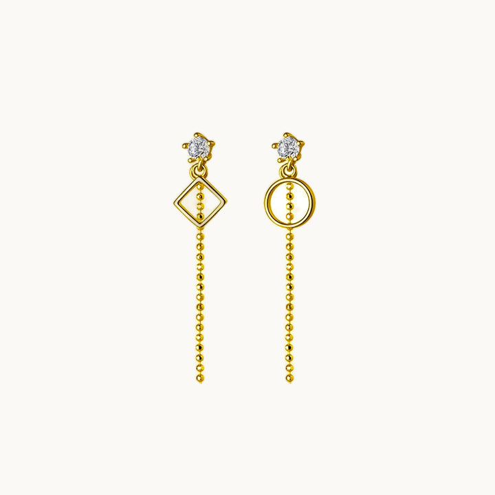 Une paire de boucles d'oreilles est exposée devant un fond beige.  Ces boucles sont composées d'un diamant sur lequel pend une chaîne et une forme géométrique qui n'est pas la même sur les deux boucles ; il y a un rond et un losange. Elles sont en argent 925 plaqué or.