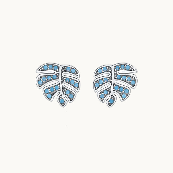 Une paire de boucles d'oreilles est exposée devant un fond beige.  Ce sont des boucles d'oreilles en forme de feuilles de palmier. Elles sont ornées de petites pierres turquoise. Elles sont en argent 925. 