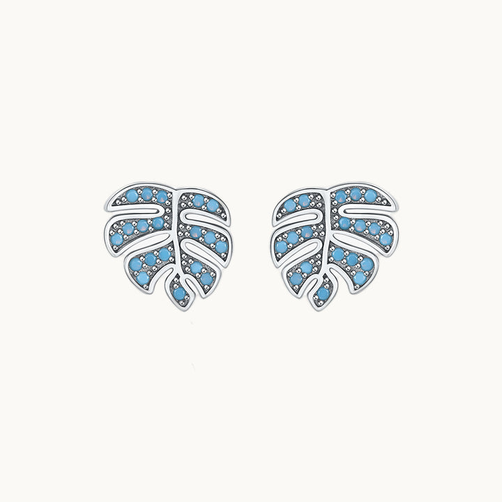 Une paire de boucles d'oreilles est exposée devant un fond beige.  Ce sont des boucles d'oreilles en forme de feuilles de palmier. Elles sont ornées de petites pierres turquoise. Elles sont en argent 925. 