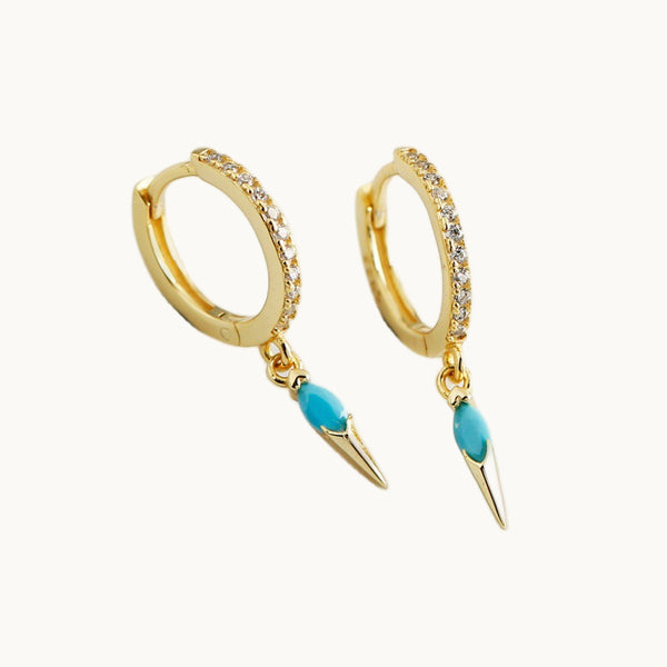 Une paire de boucles d'oreilles est exposée devant un fond beige.  Ce sont des anneaux en strass sur lesquels pend une pointe ornée d'une pierre de couleur. La boucle est plaqué or et la pierre est turquoise. 