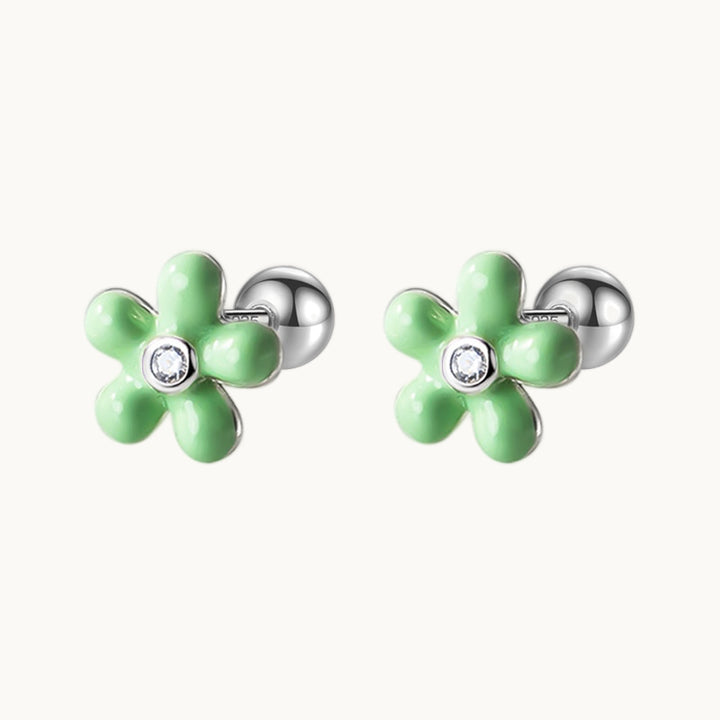 Une paire de boucles d'oreilles est exposée devant un fond beige.  Ce sont des fleurs vertes avec un strass au coeur. Les boucles sont en argent. 