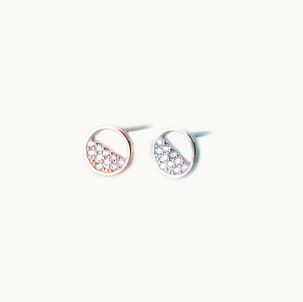 Une paire de boucles d'oreilles est exposée devant un fond beige.   Ce sont des boucles rondes en argent 925 ou en argent 925 plaqué or rose. Elles sont remplies de strass sur le bas. Les strass remplissent la moitié sur cercle de la boucle.