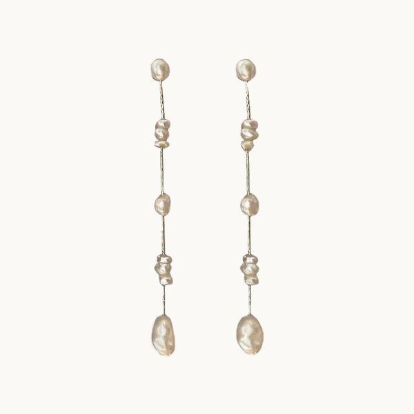 Une paire de boucles d'oreille est exposée devant un fond beige.  Ce sont des boucles d'oreille dorées  longues et pendantes. Elles sont composées d'une chaîne à maille très serrée sur laquelle pendent des perles de différentes formes et tailles. Il y a des perles rondes seules et des trios de perles plus petites aux formes plus abstraites.