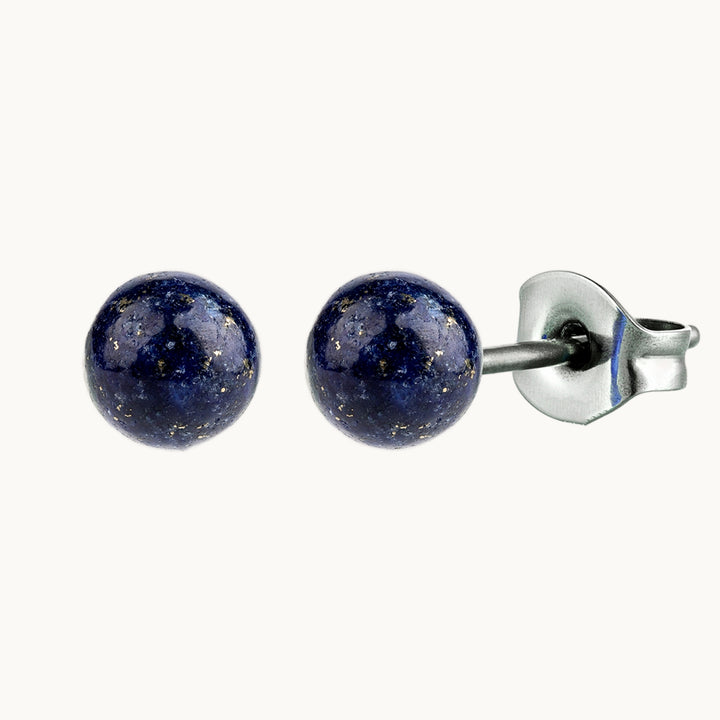 Une paire de boucles d'oreille est exposée devant un fond beige.  Ce sont des boucles d'oreille en acier chirurgical ornées d'une pierre ronde Lapis lazuli bleue. 