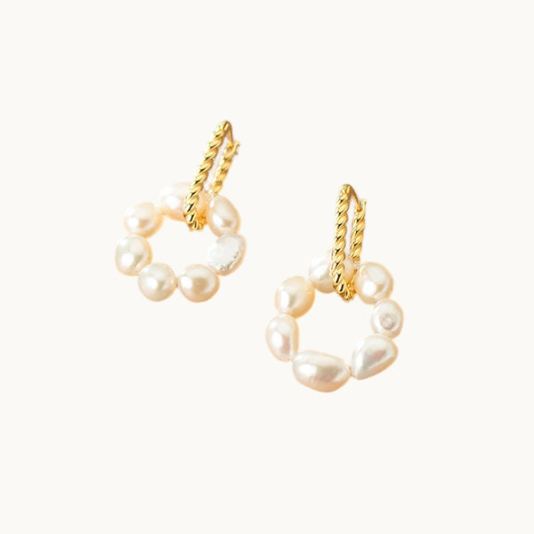 Une paire de boucles d'oreilles est exposée devant un fond beige.  Ces boucles sont composées d'un premier anneau ovale torsadé en plaqué or sur lequel est attaché un anneau ornée de perles. 