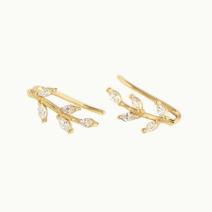 Une paire de boucles d'oreilles est exposée devant un fond beige.   Ce sont des boucles grimpantes ornées de feuilles en diamant. Les boucles sont en plaqué or. 