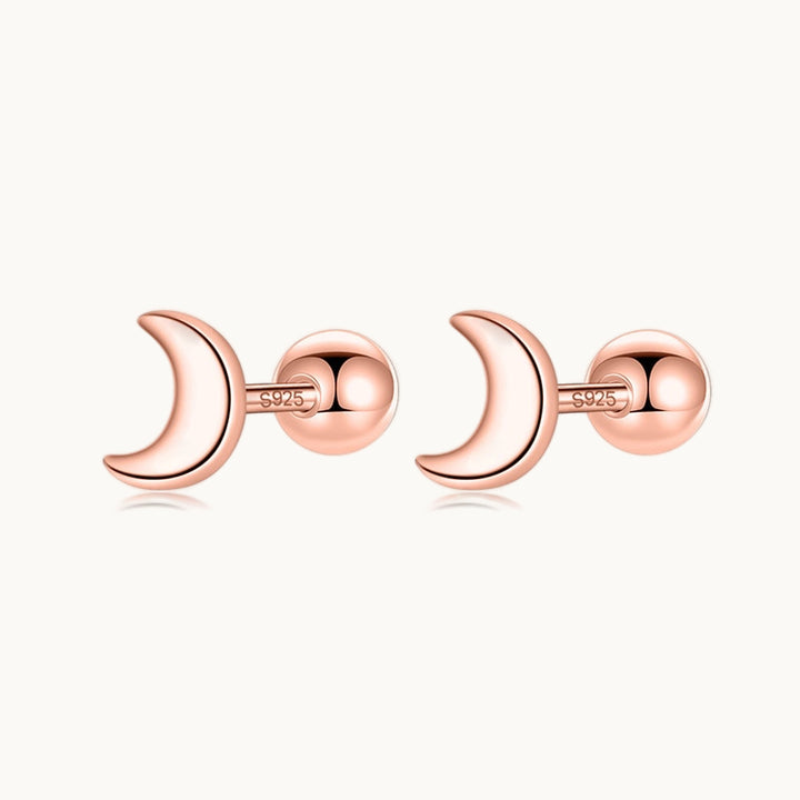 Une paire de boucles d'oreilles est exposée devant un fond beige.  Ce sont des petites lunes en or rose. 