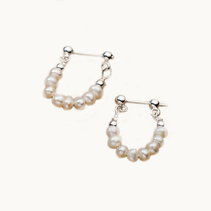 Une paire de boucles d'oreilles est exposée devant un fond beige.  Ce sont des boucles pendantes. Elles sont constituées d'une tige droite en argent  925 sur laquelle pendante des perles. Les perles font de ces boucles d'oreilles des créoles.