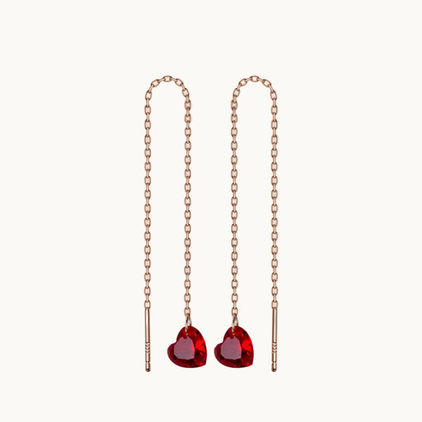 Une paire de boucles d'oreilles est exposée devant un fond beige.  Ce sont des boucles pendantes et longues. Elles sont en forme de chaîne avec un coeur en pierre de grenat rouge au bout.  Elles sont en argent 925 plaqué or rose. 