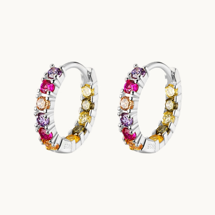 Une paire de boucles d'oreilles est exposée devant un fond beige.  Ce sont de petites créoles ornées de strass colorés  dans les tons de rose, violet et jaune. Les boucles sont en argent 925.