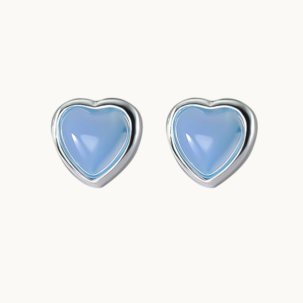 Une paire de boucles d'oreilles est exposée devant un fond beige.  Ce sont des boucles d'oreilles en forme de coeur avec une pierre transparente colorée au milieu. Elles sont en argent 925 et le coeur est bleu. 