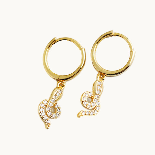 Une paire de boucles d'oreilles est exposée devant un fond beige.  Ce sont des anneaux sur lesquels pend un serpent orné de strass. Les boucles sont en plaqué or.