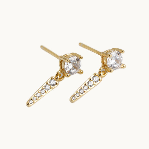 Une paire de boucles d'oreilles est exposée devant un fond beige.  Ce sont des boucles pendantes constituées d'un diamant sur lequel pend une pointe en strass. Les boucles sont en plaqué or.