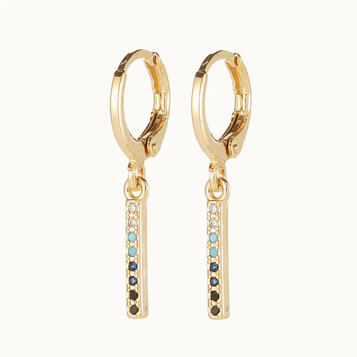 Une paire de boucles d'oreilles est exposée devant un fond beige.  Ce sont des boucles d'oreille en forme d'anneau sur lequel pend un rectangle fin orné de strass colorés. Elles sont en dorées.