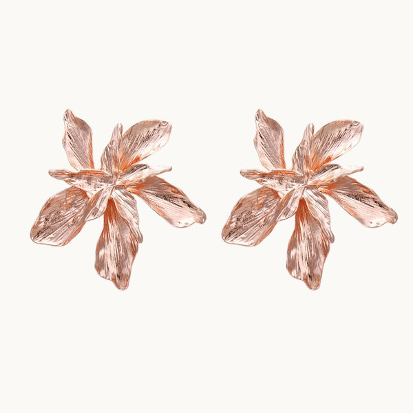 Une paire de boucles d'oreilles est exposée devant un fond beige.  Les boucles d'oreilles sont en fleurs géantes en or rose. 