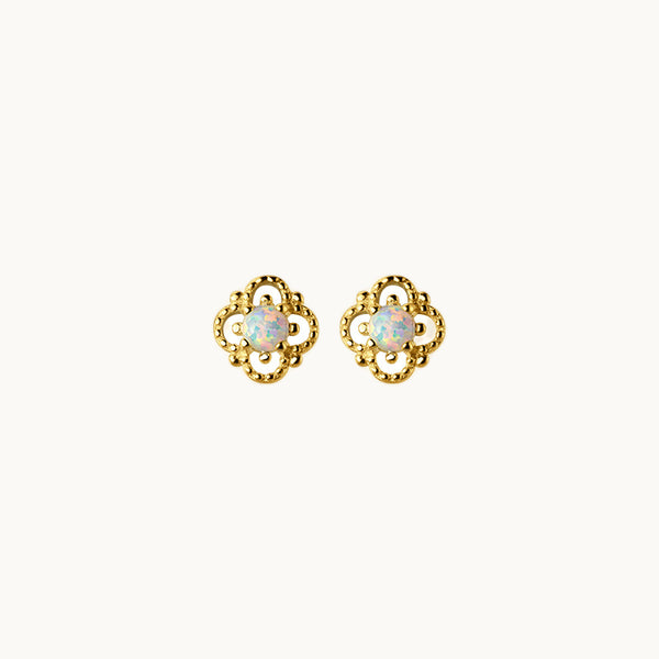 Une paire de boucles d'oreilles est exposée devant un fond beige.  Ce sont des boucles en forme de fleurs avec un strass au milieu. La forme de fleur est en argent 925 plaqué or. Le strass est de couleur blanche avec des reflets roses et bleus.