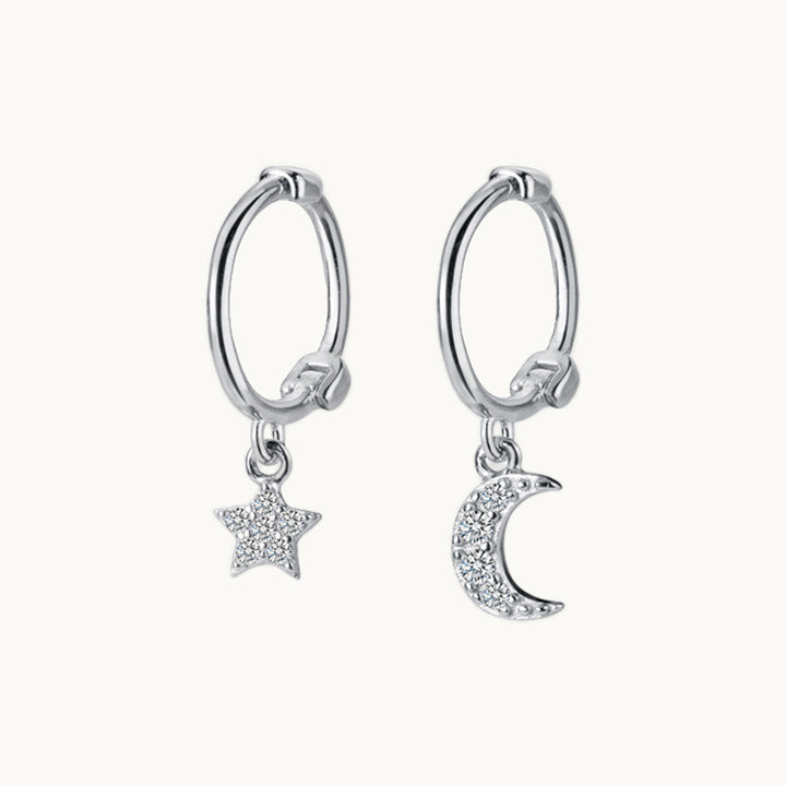 Une paire de boucles d'oreilles est exposée devant un fond beige.  Ce sont des boucles d'oreille asymétrique. Elles sont constituées d'un anneau sur lequel pend une lune et de l'autre côté une étoile. L'étoile et la lune sont ornées de strass. Elles sont en argent 925.