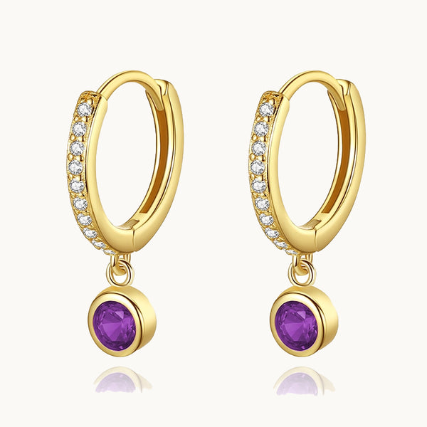 Une paire de boucles d'oreilles est exposée devant un fond beige.  Ce sont des anneaux ornées de strass sur lequel pend un diamant rond coloré. Les boucles sont en plaqué or et le diamant est violet. 