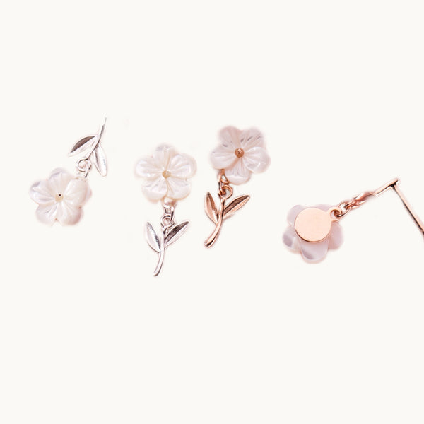 Deux paires de boucles d'oreilles sont exposées devant un fond beige.  Ce sont des boucles d'oreille en forme de fleur pendante. La tige et les feuilles sont sur le lobe tandis que le coeur et les pétales blancs de la fleur pendent.  Il y a une paire en argent 925 et une en argent 925 plaqué or rose.