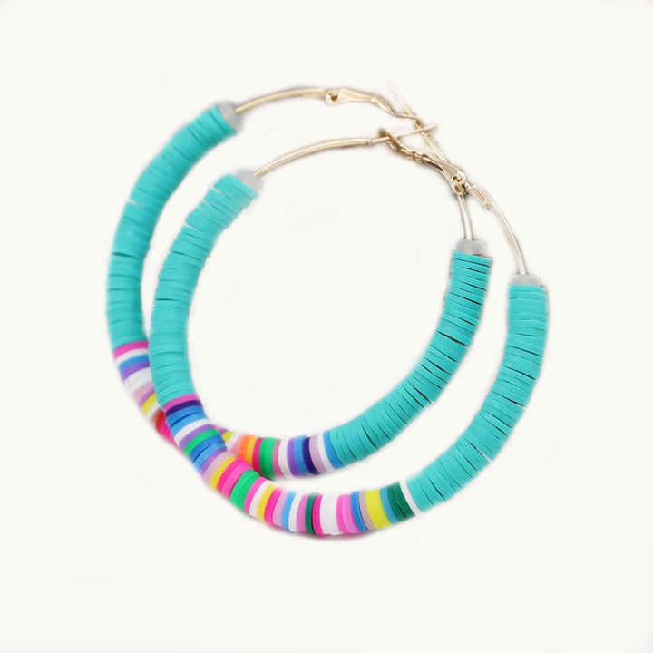 Une paire de boucles d'oreilles est exposée devant un fond beige.  Ce sont des créoles fines dorées ornées d'un tas de petits anneaux plats en argile colorés. Les anneaux sont bleu turquoise sur les extremités et le milieu des anneaux sont mulitcolores.