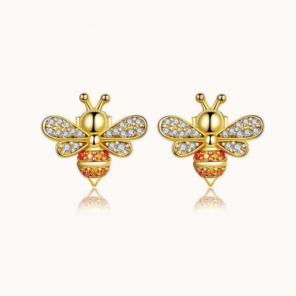 Une paire de boucles d'oreille est exposée devant un fond beige.  Ce sont des boucles en forme d'abeille dorée. Elles sont ornées de strass orange en guise de rayures et de strass transparents sur les ailes. 