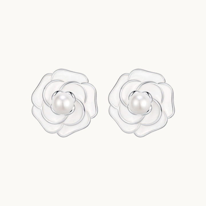 Une paire de boucles d'oreilles est exposée devant un fond beige.  Ce sont des roses blanches avec une perle au milieu. Les boucles sont en argent. 