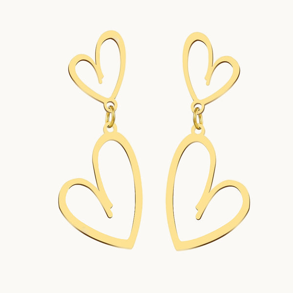 Une paire de boucles d'oreilles est exposée devant un fond beige.  Ce sont des boucles d'oreilles pendantes dorées composées de deux coeurs. 