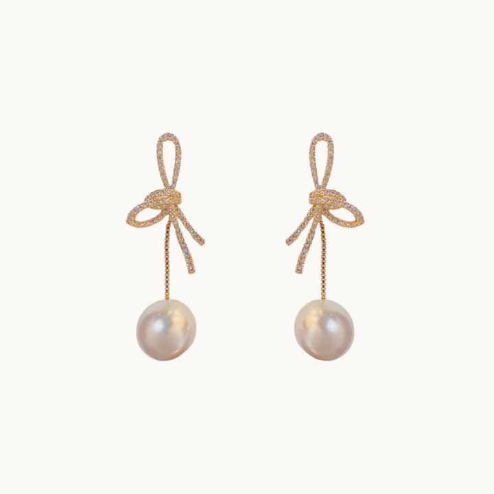 Une paire de boucles d'oreilles est exposée devant un fond beige.  Ce sont des boucles constituées d'un noeud doré avec strass sur lequel pend une perle ronde. 