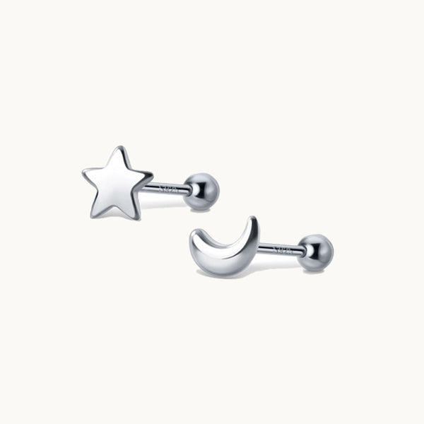 Une paire de boucles d'oreilles est exposée devant un fond beige.  Ce sont des boucles d'oreille dépareillées il y a en une lune et une étoile. Elles sont en argent. 