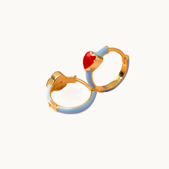 Une paire de boucles d'oreilles est exposée devant un fond beige.  Ce sont des anneaux en or et bleu orné d'un petit coeur rouge. 