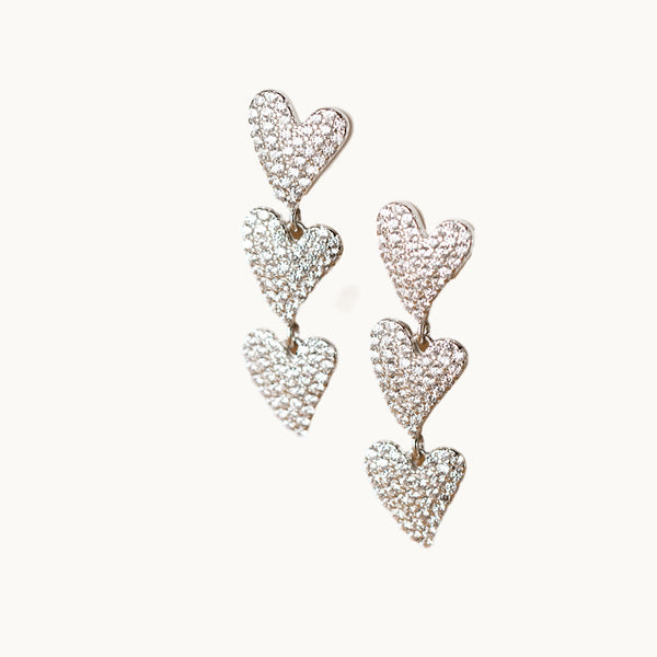 Une paire de boucles d'oreilles est exposée devant un fond beige.  Ce sont des boucles d'oreille pendantes constituées de trois coeurs ornés de strass. Elles sont en argent 925.