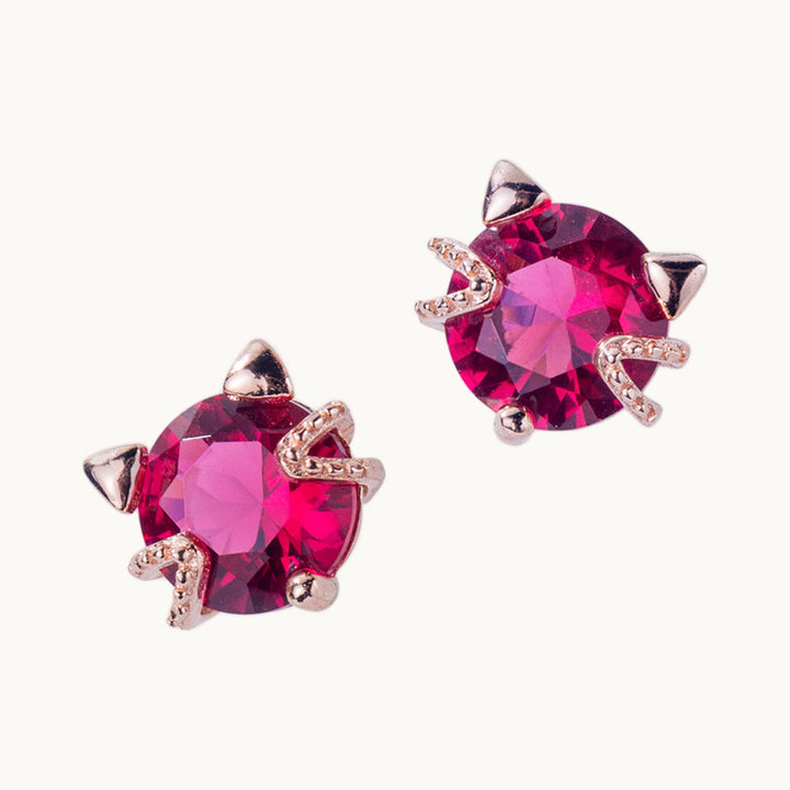 Une paire de boucles d'oreilles est exposée devant un fond beige.  Ce sont des boucles d'oreille en forme de chat. C'est une tête de chat en diamant de zirconium couleur rouge rubis sur lequel il y a des oreilles et des moustaches en argent 925 plaqué or rose.