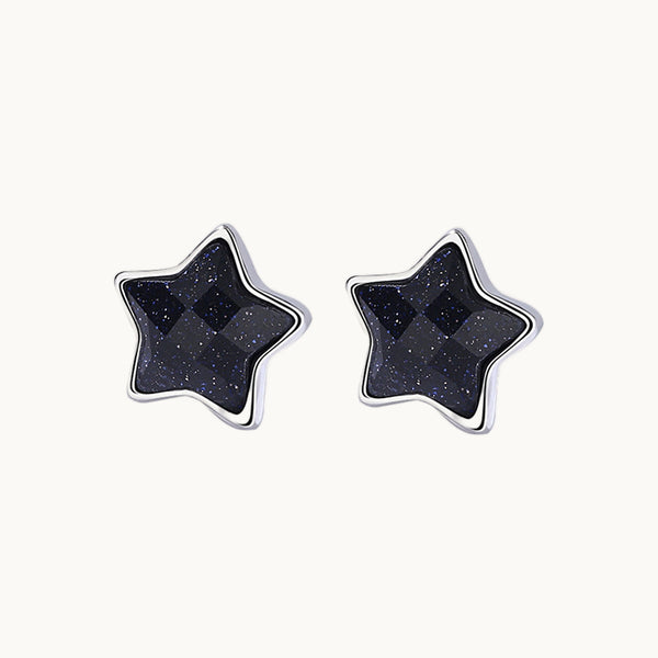 Une paire de boucles d'oreilles est exposée devant un fond beige.  Ce sont des boucles d'oreille en forme d'étoile ornée d'une pierre agate noire au milieu. Elles sont en argent 925. 