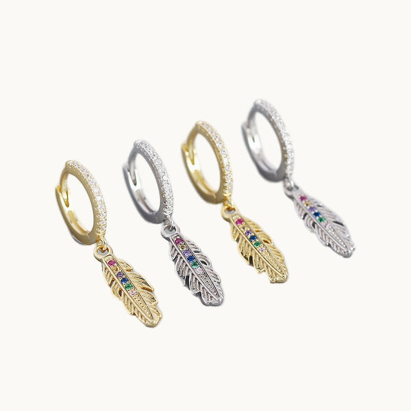 Deux paires de boucles d'oreilles sont exposées devant un fond beige.   Ce sont des anneaux ornés de strass sur lesquelles pendent une plume ornée de strass multicolore. Il y a des boucles en or et d'autres en argent. 