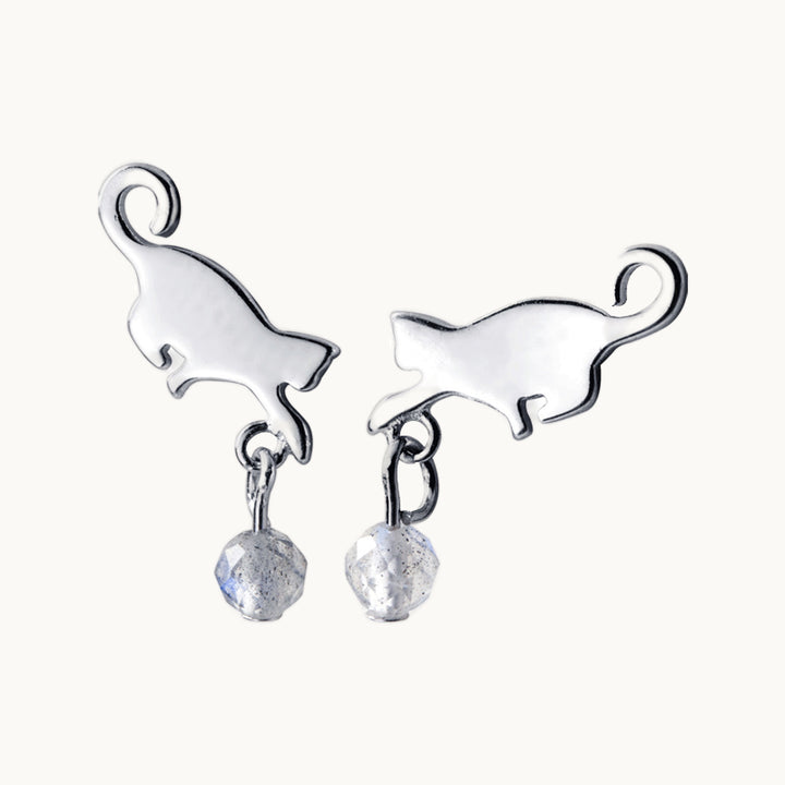 Une paire de boucles d'oreilles est exposée devant un fond beige.   Ce sont des boucles d'oreille en forme de chat de profil. Il y a une pierre de cristal blanc qui pend d'une des pattes avant du chat ce qui donne l'impression que le chat joue avec la pierre. Elles sont en argent 925.