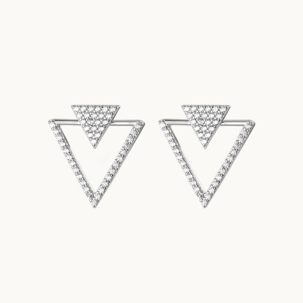 Une paire de boucles d'oreilles est exposée devant un fond beige.  Ce sont des boucles d'oreille en forme de triangles. Il y a un premier triangle plein, petit, orné de strass et un deuxième triangle beaucoup plus grand ajouré orné de strass sur deux côtés. Elles sont en argent 925. 