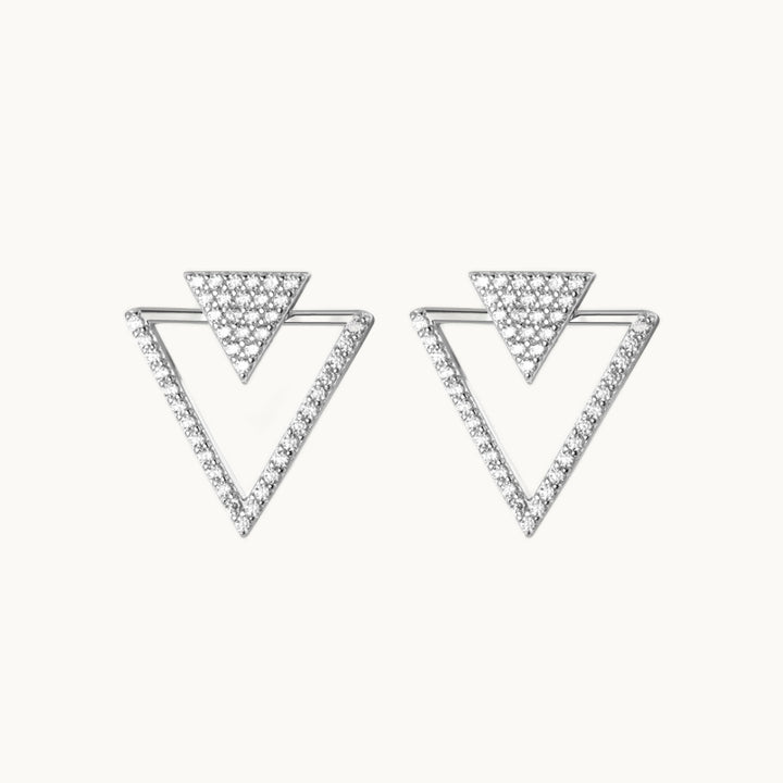 Une paire de boucles d'oreilles est exposée devant un fond beige.  Ce sont des boucles d'oreille en forme de triangles. Il y a un premier triangle plein, petit, orné de strass et un deuxième triangle beaucoup plus grand ajouré orné de strass sur deux côtés. Elles sont en argent 925. 