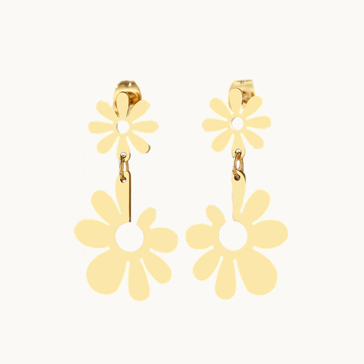 Une paire de boucles d'oreille est exposée devant un fond beige.  Ce sont des boucles d'oreille pendantes. Elles sont constituées d'une fleur sur laquelle pend une autre fleur plus grande. La petite fleur a des pétales réguliers tandis que la plus grande a des pétales de tailles variés. Elles sont en acier inoxydable plaqué or. 