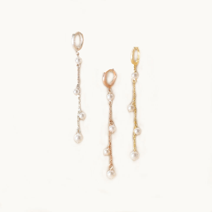 Des boucles d'oreilles sont exposées devant un fond beige.  Ce sont des anneaux sur lequel pend une chaîne ornée de perles. Il y en a une en or, une en argent et une en or rose. 