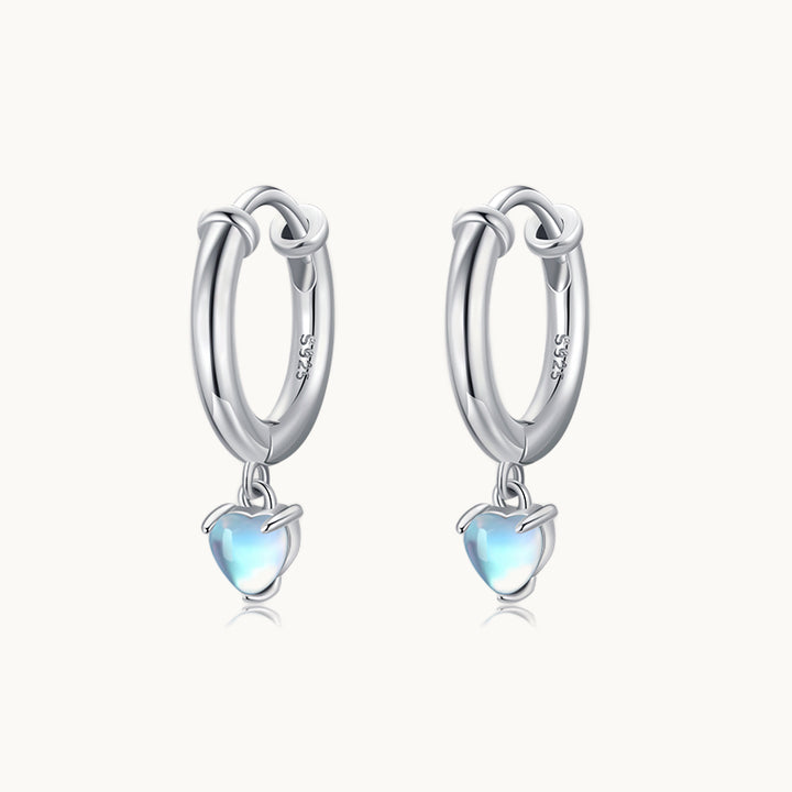 Une paire de boucles d'oreilles est exposée devant un fond beige.  Ce sont des anneaux avec un petit coeur qui pend dessus. Le coeur est en pierre de lune bleue. Elles sont en argent 925. 